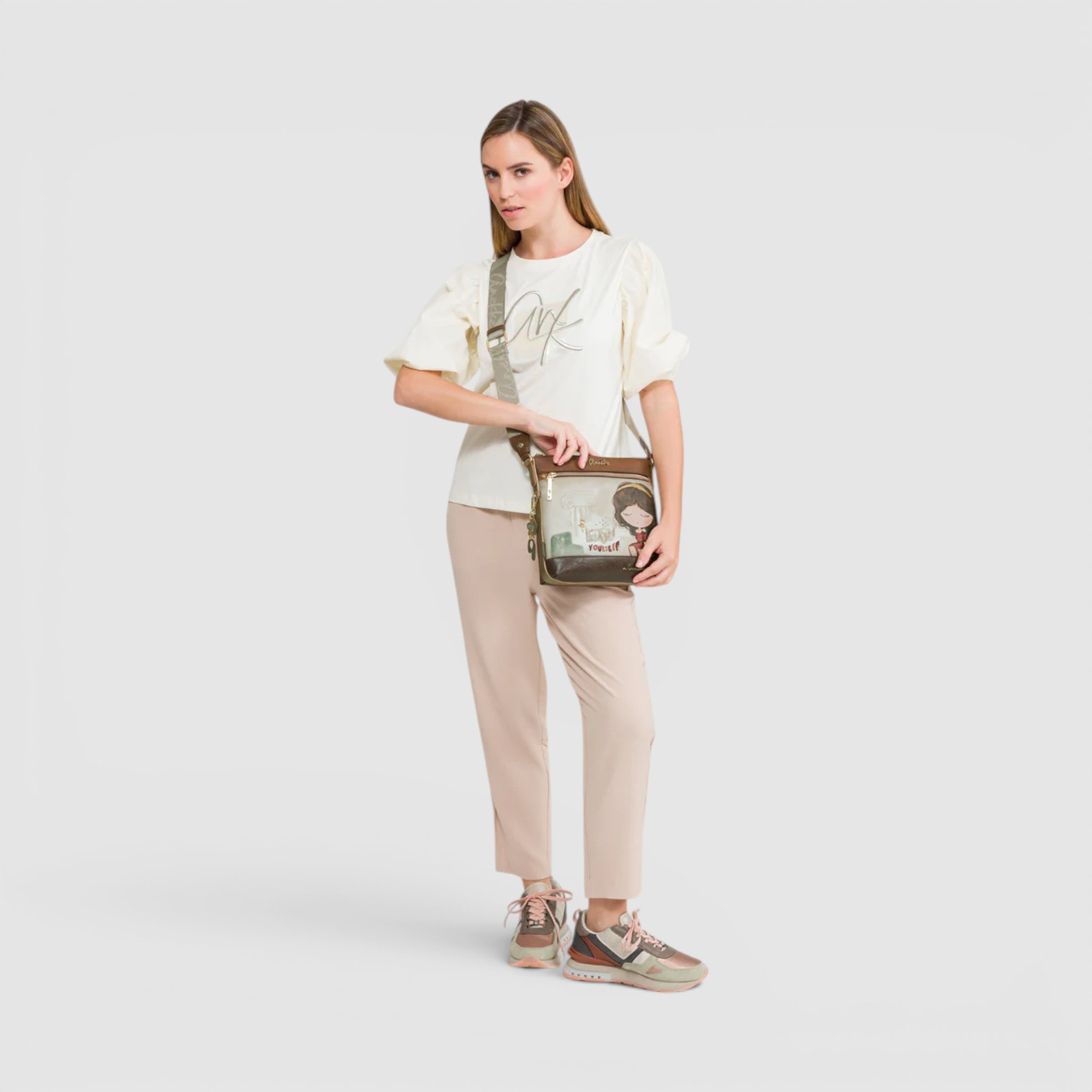 Femeie tânără în ținut casual, purtând pantaloni bej, tricou alb și pantofi sport, cu geantă de umăr.